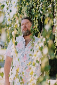 a bearded man in a floral shirt is standing in front of a weeping willow tree