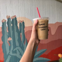 a person holding up a cup of iced coffee in front of a cactus