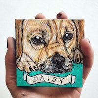 a hand holding a painting of a dog with the word daisy
