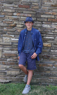 a boy leaning against a stone wall wearing a hat and shorts