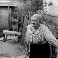 a black and white photo of an old woman standing in front of a fence