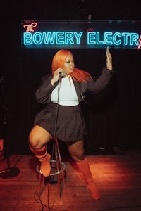 a woman with orange hair standing on a stool in front of a neon sign that says bowery electric