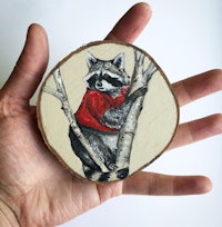 a hand holding a wooden slice with a raccoon in a red sweater