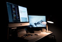 a desk with two monitors and a lamp in the dark
