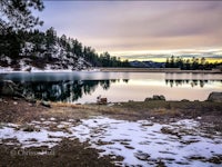 a lake surrounded by snow and trees at sunset