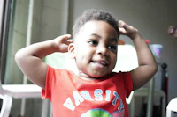 a young boy in a red t - shirt is standing in front of a chair
