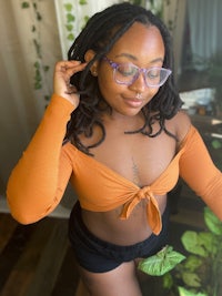 a black woman with dreadlocks and glasses is posing in front of a plant