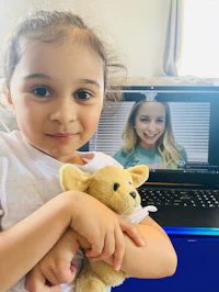 a little girl holding a teddy bear in front of a laptop