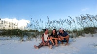 three people sitting on the sand in front of tall oleanders