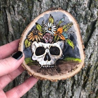 a person holding a wood slice with a skull and flowers on it
