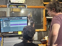 Engineer and producer looking over a console with an artist recording vocals in the live room through the glass windows separating the control room from the live room