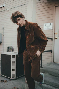 a young man in a brown corduroy jacket leaning against an air conditioner