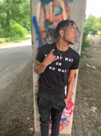 a young man leaning against a wall wearing a black t - shirt