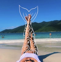 a woman laying on the beach with a mermaid tail drawn on her leg