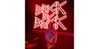 a person standing in front of a neon sign that says brick by brick