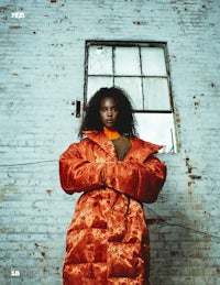 a black woman in an orange puffy coat standing in front of a brick wall
