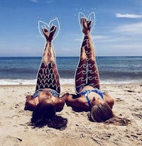 two women laying on the beach with mermaid tails drawn on their legs