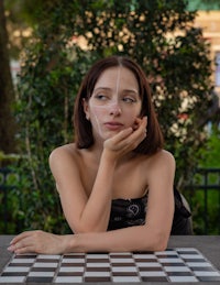a woman sitting on a chess board with a mask on her face