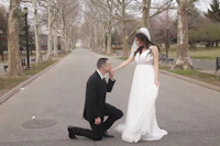 a bride and groom kneeling on a sidewalk in a park