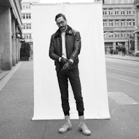 a man in a leather jacket standing in front of a white wall