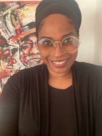 a woman wearing glasses and a turban smiles in front of a painting